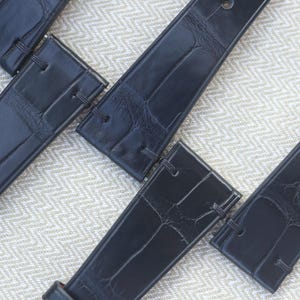 Peut inclure: Une collection de bracelets de montre noirs présentant un motif texturé en relief façon alligator. Chaque bracelet est doté de bords cousus et comprend un point de fixation à barrette à ressort en métal sur le dessus, avec une finition sombre.