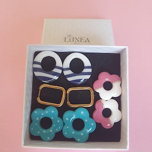 Puede incluir: Una caja blanca que contiene seis pares de pendientes. Dos pares son aros rectangulares dorados. Los otros pendientes son blancos con rayas azules, azul claro con lunares blancos y con forma de flor rosa y blanca. La caja tiene la palabra "LUNEA" impresa.