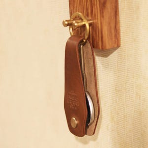 Könnte beinhalten: Ein brauner Leder-Schlüsselhalter mit einem Messingring und einem Schlüssel, der an einem hölzernen Schlüsselhaken hängt. Der Schlüsselhalter hat einen Knopfverschluss und ein geprägtes Design. Der Holzhaken ist an der Wand befestigt.