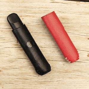 May include: A black leather case with a clear window holds a black electronic device. A red leather case sits beside it. Both cases have a textured surface and are on a light wood grain background.