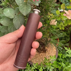 Könnte beinhalten: Eine braune Lederflasche mit silbernem Verschluss. Die Flasche ist zylindrisch und hat unten eine weiße Naht. Der Text "MAKE PEPPERS Tilly O'Goz SINCE" ist in das Leder geprägt.