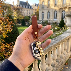 May include: A brown leather keychain with a gold-colored button clasp and a metal keyring holding car keys. The car key fob has a silver and black design with the BMW logo.