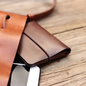 Puede incluir: Una cartera de cuero marrón con una banda elástica marrón oscuro está dentro de un bolso tote de cuero marrón. Un teléfono inteligente blanco es parcialmente visible. La bolsa y la cartera están sobre una superficie de madera.