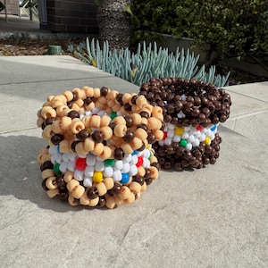 Puede incluir: Dos pulseras de cuentas hechas a mano que se asemejan a sándwiches de helado. Una pulsera es de color beige y marrón, la otra es de color marrón oscuro. Ambas tienen un centro de cuentas blancas, rojas, amarillas y verdes. Las pulseras están sobre una superficie de piedra.
