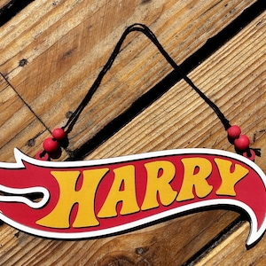 May include: A custom hanging sign featuring the name HARRY in yellow block letters on a red background with a white outline. The sign has a flame-like shape and is suspended by a black string adorned with two small red beads on each side.