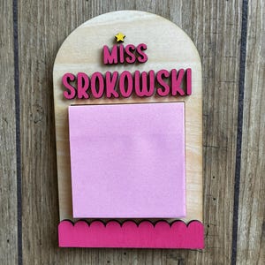 Puede incluir: Tablero de notas de madera con el nombre "MISS SROKOWSKI" en letras rosas. Un bloc de notas adhesivas rosa está adherido al tablero, que tiene un borde inferior rosa festoneado y una pequeña estrella amarilla en la parte superior.