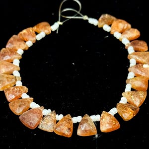 May include: A necklace featuring triangular, orange-brown sunstone beads interspersed with small, white, round beads. The sunstone beads have a sparkling, glittery appearance. The necklace is strung on a thin, clear cord.