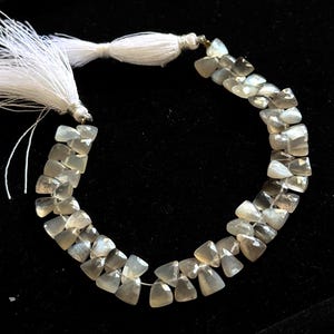 May include: A necklace of light gray, triangular-cut gemstones strung together. The necklace is accented with white tassels at each end. The gemstones have a translucent quality, and the background is black.