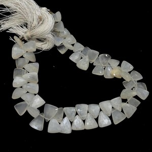 May include: A strand of pale, translucent, triangular-cut gemstones. The beads are strung together, with a white tassel at the top. The gemstones have a faceted surface, catching the light. The background is black.