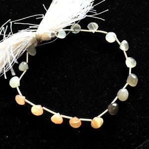 May include: A strand of gemstone beads in shades of peach, gray, and clear, strung on a silver wire. A white tassel is attached to one end. The beads are faceted and have a polished finish.