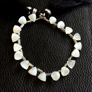 May include: A delicate bracelet featuring white, triangular and round beads strung together with silver-colored metal. The beads have a soft, translucent appearance. The bracelet is displayed against a dark, textured background.