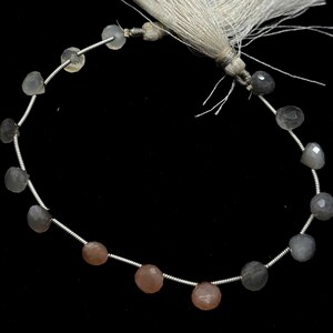 May include: A strand of faceted gemstone beads in shades of gray and peach. The beads are strung on a silver wire and are connected to a tassel. The beads have a polished, reflective surface. The background is black.