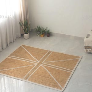 May include: A rectangular jute rug with a geometric design. The rug features a natural tan color with white lines forming a pattern reminiscent of a flag. The rug is placed on a light-colored floor, suitable for home decor.