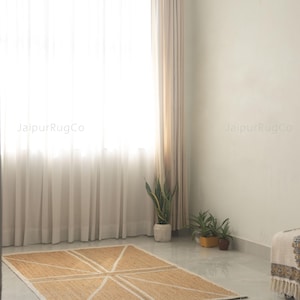 May include: A rectangular jute rug with a geometric design of white lines. The rug is placed on a glossy, light-colored floor. The room has a large window with sheer white curtains, and a few potted plants.