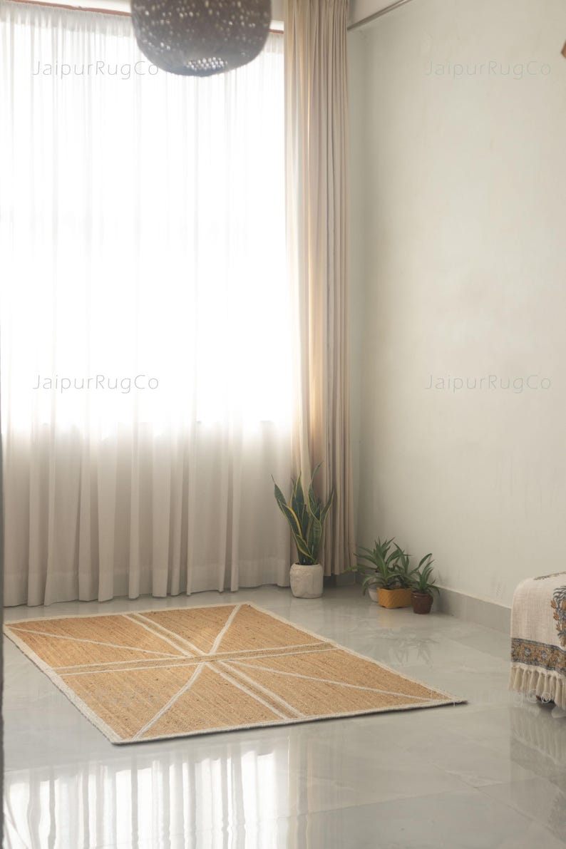May include: A rectangular jute rug with a geometric design of white lines. The rug is placed on a glossy, light-colored floor. The room has a large window with sheer white curtains, and a few potted plants.