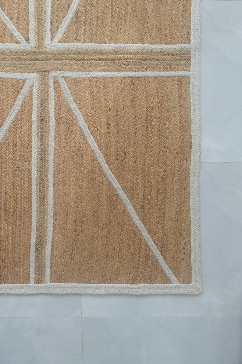 May include: A rectangular jute rug with a Union Jack design. The rug features a natural tan background with a white and tan geometric pattern. The border is white, and the rug is placed on a light gray surface.