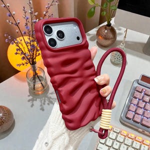 May include: A burgundy phone case with a textured, wavy design. The case has a matching burgundy wrist strap with a beige accent. The phone case is designed to fit a smartphone with multiple camera lenses.
