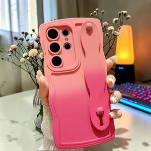 Könnte beinhalten: Eine pink-magentafarbene Ombre-Handyhülle mit einem Riemen. Die Hülle hat ein strukturiertes Design und Aussparungen für die Kameralinsen des Telefons. Der Riemen hat ein kleines blütenförmiges Detail am Ende.