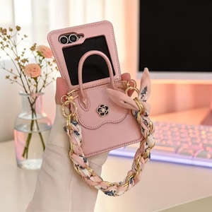 Può includere: Una custodia per telefono rosa chiaro progettata come una borsa in miniatura con manico superiore e chiusura a patta. Presenta un piccolo emblema a forma di fiore bianco e nero e una catena dorata intrecciata con un nastro in tessuto rosa.