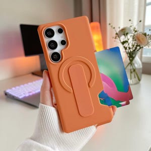 Könnte beinhalten: Eine matte orangefarbene Smartphone-Hülle mit integriertem vertikalen Ständer und einem Kartenfach auf der Rückseite. Die Hülle hat einen Ausschnitt für eine Kamera mit mehreren Linsen und enthält eine rechteckige Karte mit einem farbenfrohen, abstrakten Farbverlauf in Rosa, Blau und Grün.