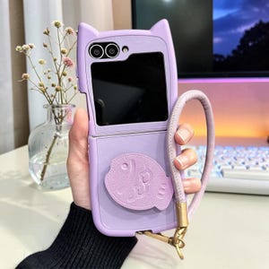 May include: A lilac phone case featuring cat ears and a fish motif. The case is open, displaying the phone's screen. A matching lilac wrist strap is connected with a gold-coloured clasp. The phone case is designed to protect the phone.