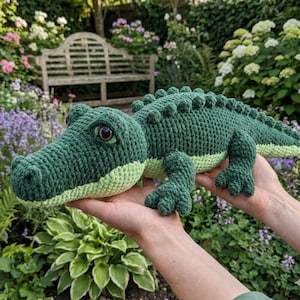 May include: A handmade crochet alligator toy featuring a dark green textured back with small raised bumps and a light green underbelly. The stuffed animal has a long snout, a thick tail, and dark brown plastic eyes, crafted with yarn.