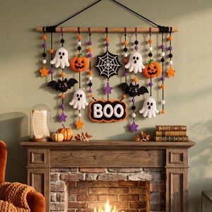 Op de afbeelding: Een wandhanger met gehaakte Halloween-figuren, waaronder witte spoken, oranje pompoenen, zwarte vleermuizen, een spinnenweb en een bordje met de tekst BOO, opgehangen aan een houten stok met zwarte koorden en oranje, paarse, witte en zwarte kralen.