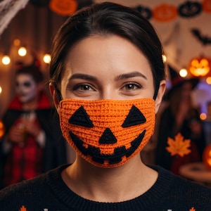 May include: A bright orange crochet face mask featuring a black jack-o'-lantern face design with two triangular eyes, a triangular nose, and a jagged, toothy grin. The mask is secured with black elastic ear loops and has a textured yarn finish.