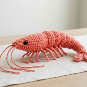 May include: A handmade crochet shrimp in a coral color. The shrimp has a segmented body, multiple legs, and long, thin antennae. The eyes are small and black. The shrimp is lying on a white surface.