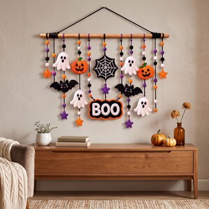 Op de afbeelding: Een Halloween-wandhanger met een houten stok en zwarte koorden waaraan gehaakte figuren hangen, waaronder witte spoken, oranje pompoenen, zwarte vleermuizen, een spinnenweb, paarse en oranje sterren en een bordje met de tekst BOO in het wit.
