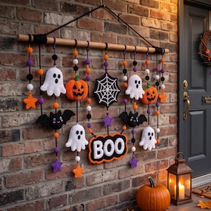 Op de afbeelding: Een Halloween-wandhanger met een houten stok, zwart touw en diverse gehaakte ornamenten, waaronder witte spoken, oranje pompoenen, zwarte vleermuizen, een wit spinnenweb en een bordje met het woord BOO, versierd met kleurrijke kralen.