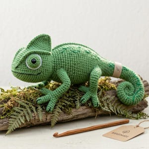 May include: A handmade, green crochet chameleon figurine with a curled tail, perched on a moss-covered log. The chameleon has a detailed, textured design and a large, round eye. A wooden crochet hook and a "HANDMADE" tag are also visible.