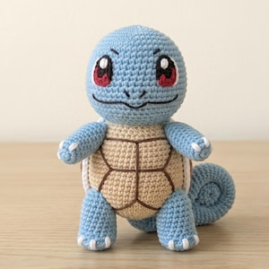 May include: A light blue crocheted turtle-like creature with a tan shell featuring dark brown stitched lines. The figure has large eyes with red irises, black pupils, and white highlights, a small mouth, and a curled tail, all crafted from yarn.