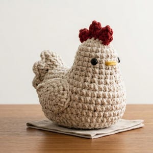 Puede incluir: Una figura de gallina hecha a mano con ganchillo en hilo beige, con cresta roja y ojos negros. La gallina está sobre un pequeño paño cuadrado beige. El pico es amarillo. La figura está sobre una superficie de madera.