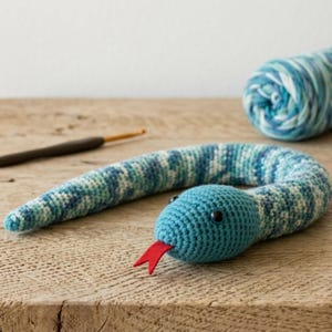 Op de afbeelding: Een handgemaakt gehaakt slangenknuffel in blauwe, groene en witte tinten, met een felrode tong. De kop van de slang is effen turquoise blauw en heeft zwarte ogen. Een bol wol en een haaknaald zijn op de achtergrond te zien.