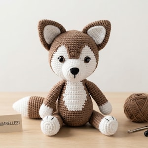 May include: A handmade crochet fox toy in brown and white. The fox has large, upright ears, a white chest, and a fluffy tail. It is sitting on a wooden surface. The fox is a soft toy.