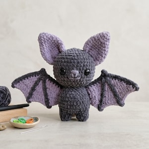 May include: A handmade crochet bat in shades of gray and purple. The plush toy has large ears and wings, with black eyes and a small nose. The bat is standing upright, showcasing its detailed craftsmanship.