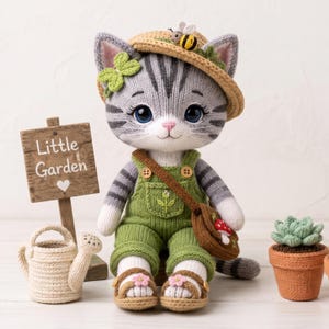 Könnte beinhalten: Eine gehäkelte graue getigerte Katzenpuppe mit Strohhut und Biene, grüner Latzhose und Sandalen. Die Katze trägt eine braune Umhängetasche mit Pilz, ein grünes Kleeblatt am Ohr und steht neben einem Holzschild mit der Aufschrift Little Garden.