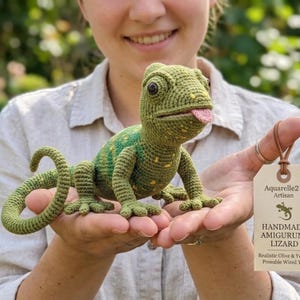 May include: A handmade amigurumi lizard in shades of green and yellow, with a pink tongue sticking out. The crochet lizard has a realistic design and poseable wired tail. The tag reads "Aquarelle2 Artisan".