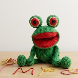 May include: A handmade green crochet frog with large, round eyes and a wide red mouth. The frog is sitting on a wooden surface, with colorful yarn scraps scattered around. The frog's eyes have a white and black design.