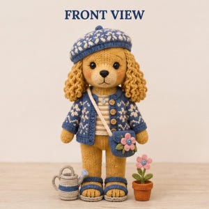 Può includere: Un orsacchiotto all'uncinetto con pelliccia dorata, che indossa un basco blu a motivi, un cardigan blu abbinato con disegni di fiocchi di neve bianchi e una maglietta a righe. Porta una borsa a tracolla blu con un fiore rosa e un piccolo annaffiatoio.