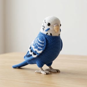 以下が含まれることがあります： 手作りの青と白のかぎ針編みのセキセイインコ。鳥は、黒い模様のある白い頭、黄色のくちばし、白いディテールのある青い翼を持っています。ベージュのかぎ針編みの足の上に立っており、木の表面に置かれています。