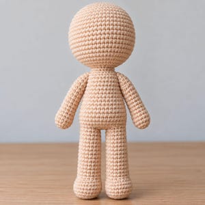 Puede incluir: Una base de muñeco de ganchillo en color beige con una cabeza grande y redonda, un torso cilíndrico, dos brazos y dos piernas. La figura está hecha con un patrón de puntos apretados y uniformes en hilo color piel, creando una textura lisa.