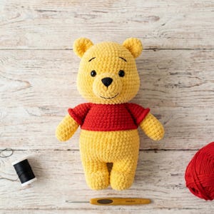 May include: A yellow and red crocheted bear plush toy with black embroidered eyes and nose. The bear wears a red shirt and has a textured yarn surface. The toy is positioned with a spool of black thread, a wooden crochet hook, and red yarn.