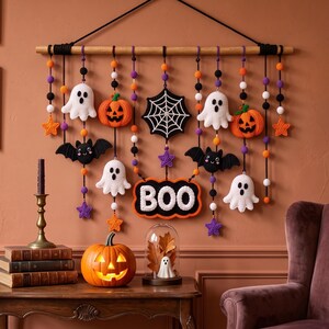 Op de afbeelding: Een Halloween-wandhanger met gehaakte spoken, pompoenen, vleermuizen, een spinnenweb en een bordje met de tekst BOO. De decoraties hangen aan een houten stok met zwarte koorden die zijn versierd met oranje, paarse, witte en zwarte kralen.