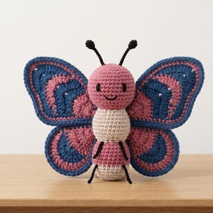 May include: A crocheted butterfly toy featuring a pink head, a beige and pink striped body, and large wings with concentric layers of blue and pink yarn. The butterfly has two black antennae and black embroidered eyes and a small curved mouth.