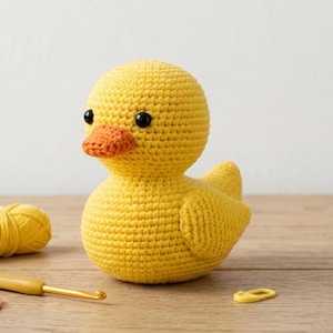 Puede incluir: Un pato amarillo tejido a mano con un pico naranja y ojos negros. El pato está sobre una superficie de madera. También se ven un ganchillo, hilo y marcador de puntada amarillos.