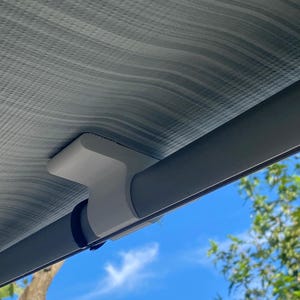 以下が含まれることがあります： グレーと白のストライプの日よけの一部で、灰色の金属製のポールで支えられています。白いプラスチック製のブラケットが日よけをポールに接続しています。背景には青い空と緑の葉が見えます。