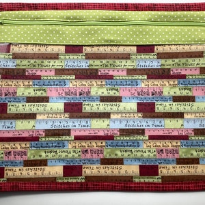 May include: A rectangular quilted sewing project bag with a red patterned border. The main body features a ruler design in various colors, with text like "Stitches in Time" and "Bear Paw Shoppe." A green polka-dot fabric panel with a zipper closure is at the top.