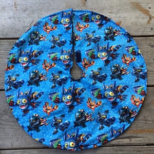May include: A blue circular Christmas tree skirt with a repeating pattern of cartoon characters. The characters are blue, orange, green, and black. The characters are on a blue background with white dots.